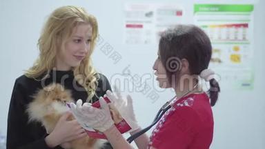 一位美丽的女士，怀里抱着一只小狗，兽医在一家兽医诊所用听诊器给她画像