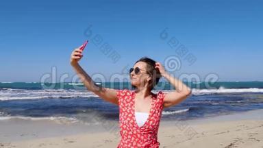 年轻的美女穿着红色连衣裙，带着相机手机在海风和海浪的海滩上自拍。 慢动作