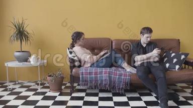 年轻夫妇坐在家里的沙发上，<strong>沉迷</strong>于现代小玩意。 库存录像。 使用手机的人