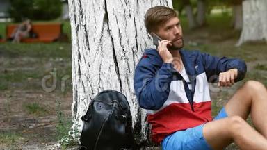 一个年轻人谈论用他的智能手机开会，<strong>坐在</strong>公园的一棵<strong>树下</strong>。 使用移动电话的蓄须学生