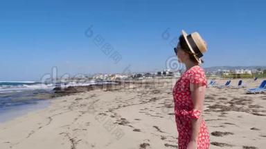 年轻漂亮的女人，穿着红色的连衣裙和帽子，独自站在空荡荡的海滩上，躺着日光浴。 沙滩，有强烈的波浪和风。