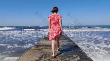 年轻女子赤着脚走在海上码头上，而海浪<strong>撞<strong>击</strong>码头。 <strong>后</strong>面的镜头。 慢动作