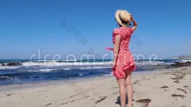 年轻迷人的女人拿着她的帽子，而红色的裙子正在大风中拍打。 她站在空荡荡的海滩上看着拖