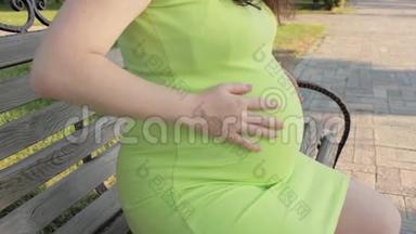 一位孕妇坐在公园的长凳上，从下到上抚摸她的腹部。 怀孕期间在大自然中行走。 等待
