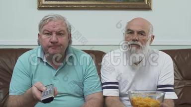 两个成熟的高级男人坐在棕色皮革沙发上看电视的肖像。 一个拿着薯片的人