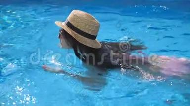 年轻漂亮的女孩戴着太阳镜和帽子在游泳池游泳。 在阳光明媚的日子里，女人在清澈的温水中放松。 夏季