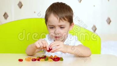 小快乐的孩子坐在桌子旁，看着明亮的水果糖果。 男孩拿着糖果吃了它