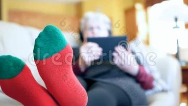 穿着红袜子的老妇人在家里的沙发上用平板电脑和耳机放松。 祖母阅读和听音乐。