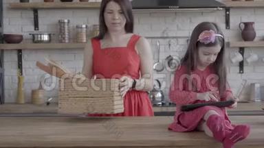 穿着粉红色连衣裙的小女孩坐在桌子上玩平板电脑。 妈妈把带面包的柳条篮子放在桌子上