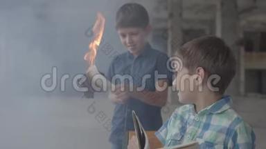 两个小双胞胎兄弟在烟雾弥漫的房间里。 一个男孩用打火机点燃了报纸，另一个孩子在看书