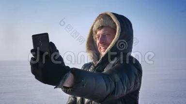 穿着<strong>保暖</strong>外套和引擎盖的商人在雪地沙漠的智能手机上有一个视频卡的特写肖像。