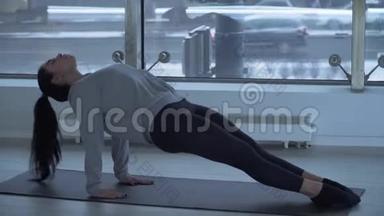 年轻的黑发女人抬起她的身体，伸展双腿坐在健身房的垫子上，上面有大的<strong>落地</strong>窗