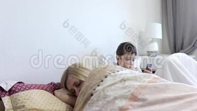 疲惫的母亲睡觉和她的太阳坐在<strong>床上</strong>，<strong>玩手机</strong>游戏的小工具，智能<strong>手机</strong>。 缺乏关注