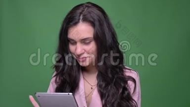 身着<strong>粉色</strong>夹克的漂亮黑发女商人用平板电脑对着镜头，在绿色的背景上羞涩地<strong>微</strong>笑