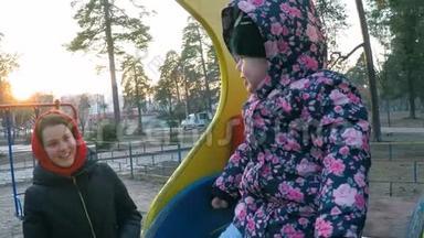 一个漂亮的小女孩坐在她母亲旁边，穿着一件五颜六色的夹克，坐在春天公园的山上，上面有高高的松树。