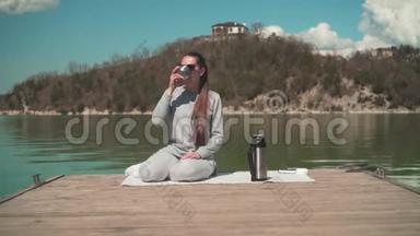 一个女人在阳光下喝热水瓶里的茶，在春天的一天坐在湖边的一个木墩上，在大自然中放松。