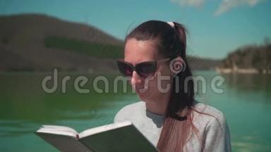 一个年轻的女人正在阳光下阅读一本书，在春天的一天，坐在一个湖的<strong>木墩</strong>上，在大自然中放松。