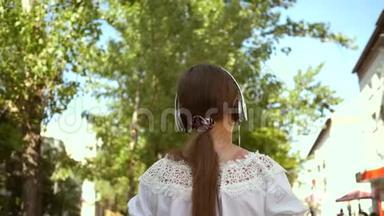 快乐的女孩戴着耳机听着<strong>音</strong>乐走在城市的<strong>街道</strong>上。 穿着白色长裙长发的女孩