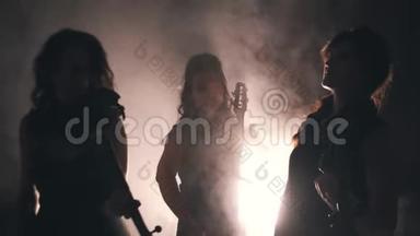 三个伟大的女孩小提琴手在一个烟雾弥漫的房间里演奏。 光线从烟雾中穿过. 很漂亮的灯光。
