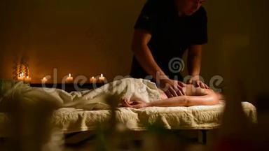 特写水疗按摩妇女`肩膀和背部。 男人的手在黑暗的房间里给女人按摩，里面有蜡烛