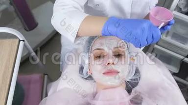美容师用刷子把白色`面具戴在女人的脸上。 戴着蓝色橡胶手套的美容师的手。 面部