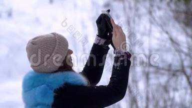 年轻漂亮的女孩走在白雪皑皑的冬林里。 她`高兴。 一个女孩正在智能手机上拍照。 做一个
