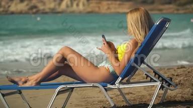年轻的金发<strong>女人</strong>在海滩上<strong>晒太阳</strong>，躺在甲板椅上，背景是大海。 女孩在手机上发短信