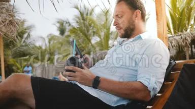 成功的快乐男游客，带着智能手表和水果饮料，坐在沙滩椅上，使用智能手机购物应用程序。