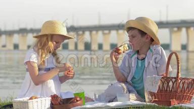 野餐的小朋友，饥饿的男孩和女孩在河边的大自然野餐时吃甜面包和喝牛奶