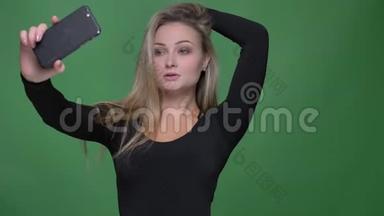年轻模特穿黑色衬衫的肖像，在绿色背景下使用智能手机制作自拍照片。