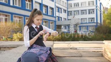 十几岁的女孩坐在学院附近的栏杆上，背着一个背包，看着他的智能手表。