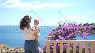从露台上看到美丽的海景，有一棵开花的树。 妈妈和宝宝欣赏海天相接，假期与