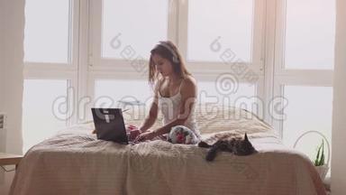 年轻漂亮的女人穿着睡衣坐在窗边的床上。 与笔记本电脑合作。 缅因州的猫坐在附近
