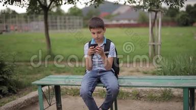 小男孩背着背包坐在长凳上，课间玩网络游戏