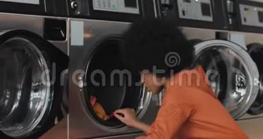 年轻的非洲裔美国妇女坐在洗衣机前，拿走洗衣服。 自助服务公众