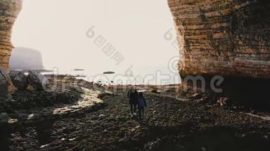 在史诗般巨大的岩石拱门诺曼底下，幸福的游客男女沿着令人惊叹的卵石海滩漫步的空中后景。