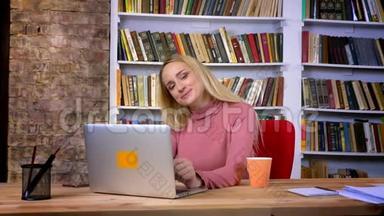 白种人年轻女孩的肖像，穿着刺，坐在笔记本电脑前，害羞地摆姿势，在书架上微笑