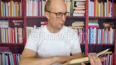 体贴的白人在图书馆内部的书架背景下读书。 戴着眼镜和白色t恤的男人坐在