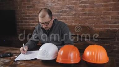 男领班坐在工地办公室的办公桌后面，重新计算头盔。
