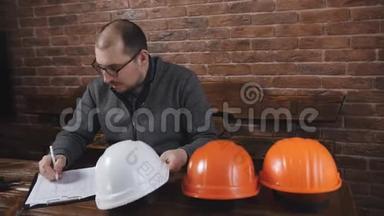男领班坐在工地办公室的办公桌后面，重新计算头盔。