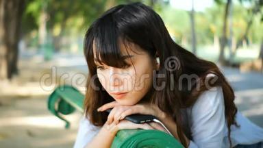 美丽的年轻时髦女郎悲伤的亚洲女人带着智能手机带着耳机听音乐，坐在公园的长凳上。