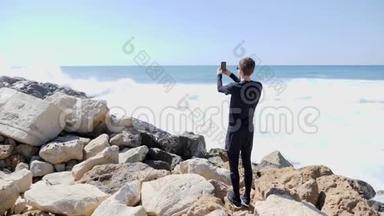 一个年轻的白种人站在岩石海滩上，手里拿着电话，一边说着话，一边拍着海浪拍打岩石的照片