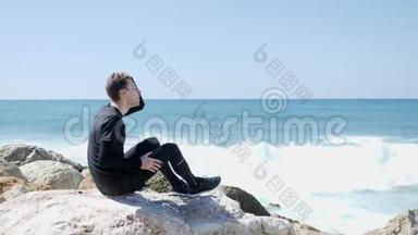 年轻的白种人坐在岩石上，手指插在头发上。强烈的海浪拍打着溅起水花的岩石海滩