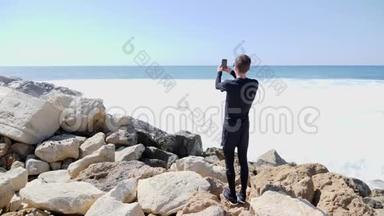 一个年轻的白种人站在<strong>岩石</strong>海滩上，手里拿着电话，一边说着话，一边拍着<strong>海浪</strong>拍打<strong>岩石</strong>的照片