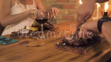 男人和女人一边切肉牛排，一边在晚上的餐厅用蜡烛吃饭。 浪漫的情侣在烧烤餐厅用餐