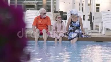 有趣快乐的成熟夫妇和小孙女坐在豪华游泳池的边缘。 祖母、祖父和