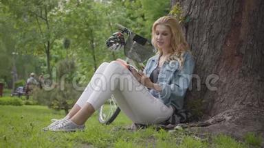 肖像可爱的微笑的女人坐在公园的一棵老树下大声朗读日记。 现代自行车站着