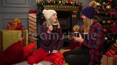 两个穿着红色格子衬衫的女孩坐在圣诞树下，戴着温暖的白色和紫色的帽子，谈论她们的事情。
