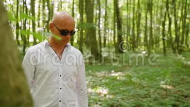 一个戴太阳镜的秃头穿过树林的肖像。 绿树，夏天，森林，大自然.. 中弹。