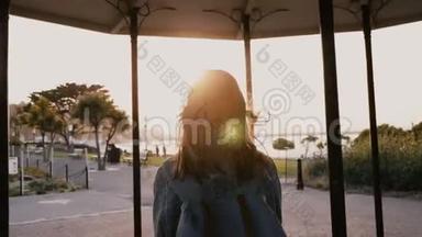 镜头跟随快乐休闲的年轻女子背着背包，头发随风飘散，沿着夏日夕阳西下的海滨<strong>公园漫步</strong>